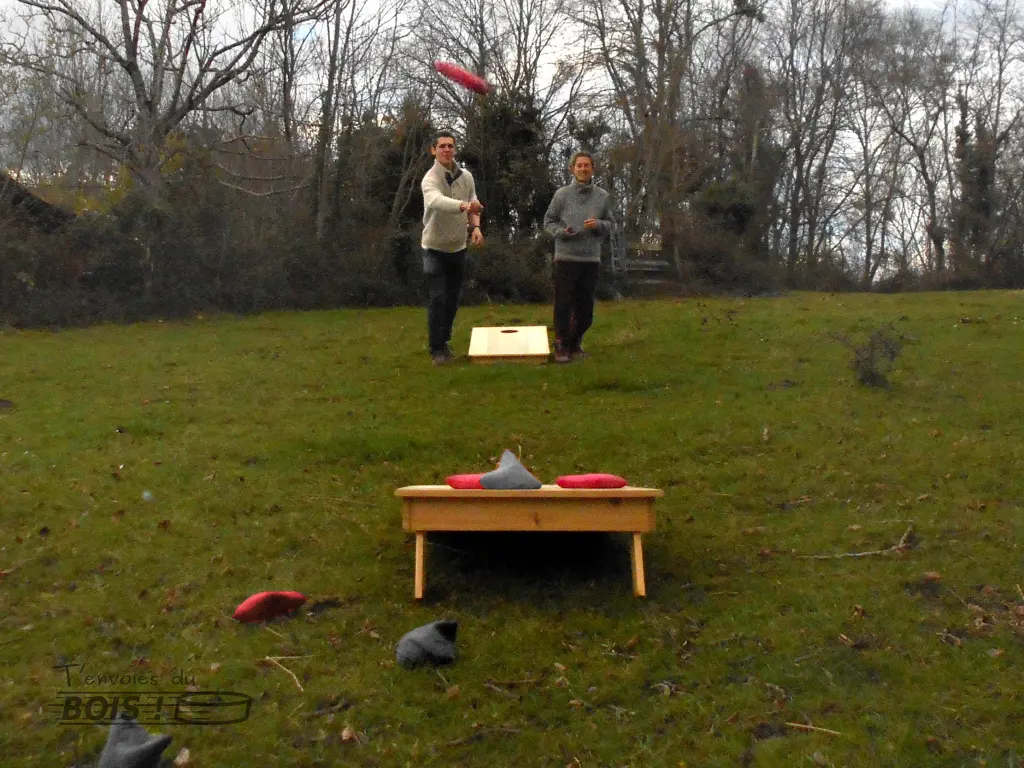 Deux personnes jouent au cornhole. Un sac de maïs pris en plein vol, en bonne voie pour rentrer dans le trou.