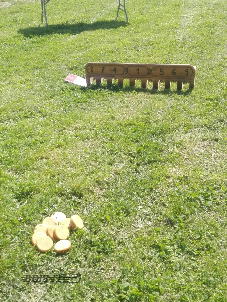 Un jeu de trou madame posé sur l'herbe avec, à quelque distance, ses dix galets en bois. Il est prêt à jouer.