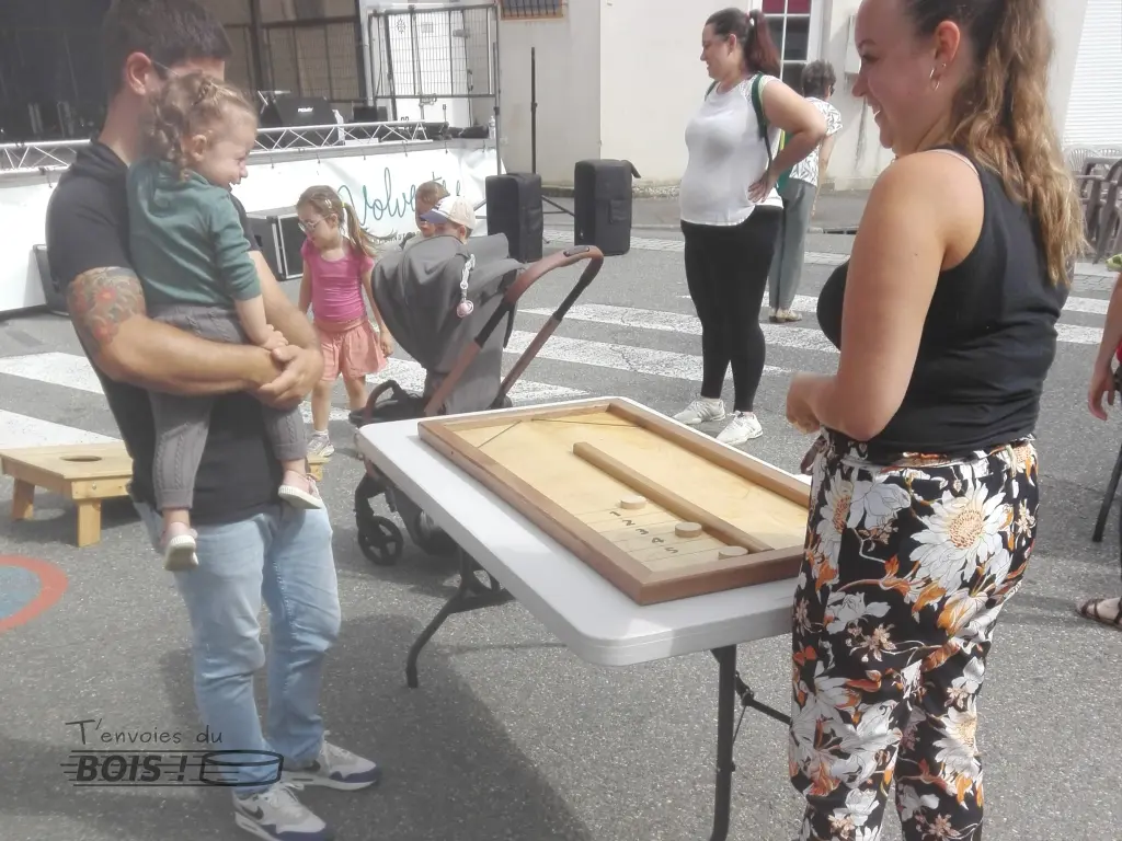 Un enfant, dans les bras de son père, est émerveillée par la partie de billard à rebonds que joue sa mère. Scène capturée lors de la fête de Mauzac en 2025.
