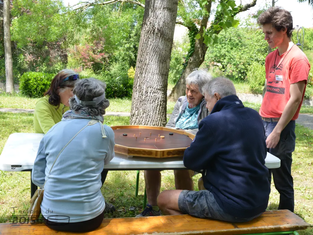 Quatre cyclistes de passage jouent ensemble au crokinole sous la supervision de votre serviteur, Alexandre Sahuguède. Scène capturée lors d'une après-midi jeux en bois à Montesquieu-Volvestre en 2025.