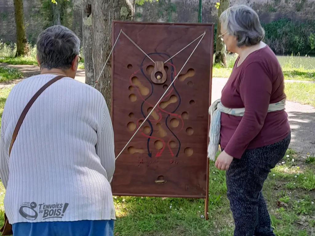 Deux personnes âgées jouent au slalom géant, la première contrôle la nacelle avec les anneaux, la seconde observe et conseille. Photo capturée lors de l'après-midi jeux en bois à Montesquieu-Volvestre en 2024.
